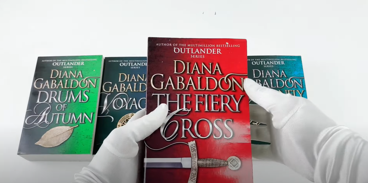 A hand wearing gloves holding Diana Gabaldon's book, with other books arranged below